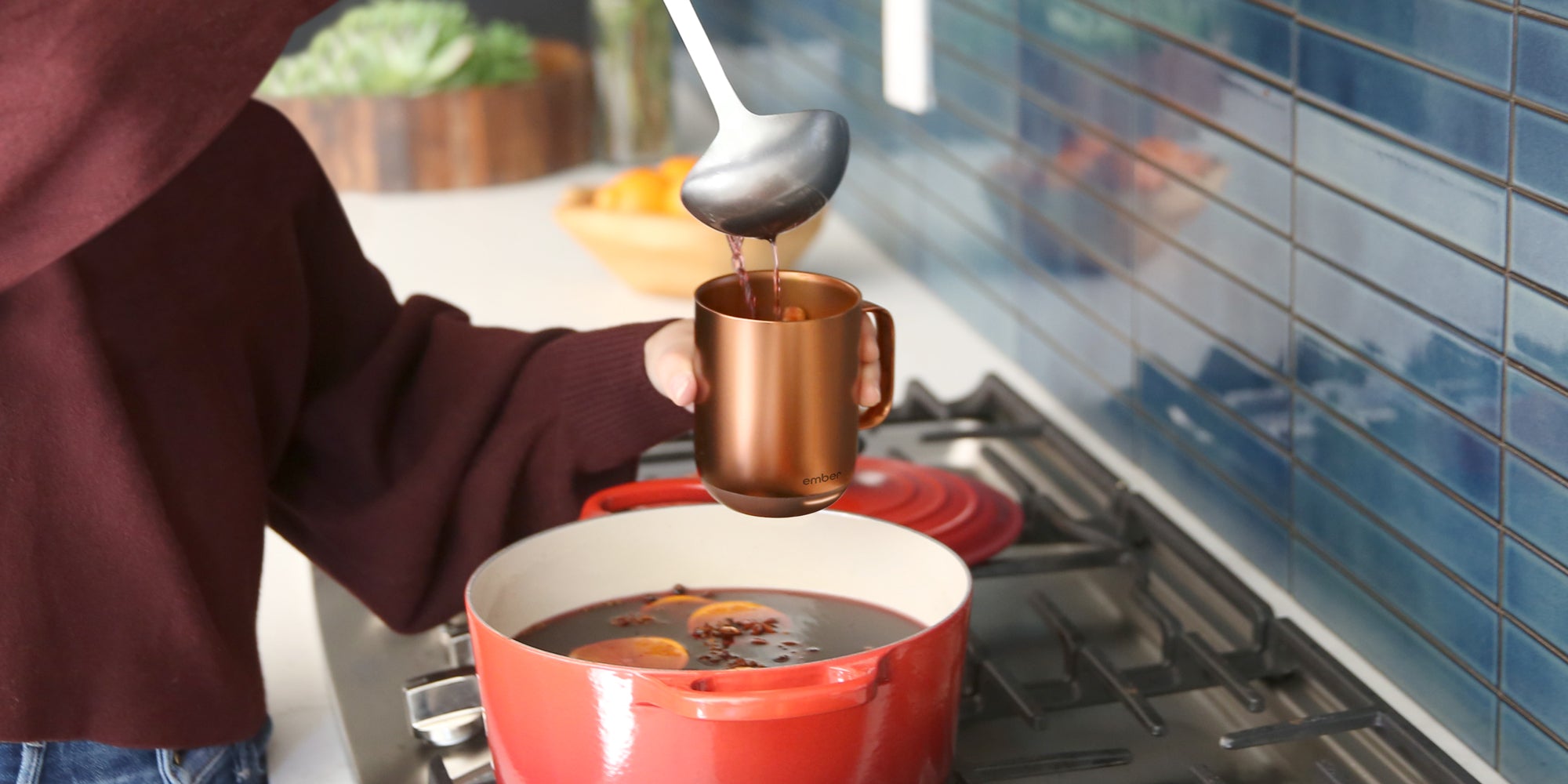A person holding an Ember Mug² Copper Edition over a red pot of mulled wine while they ladle the beverage into the mug.
