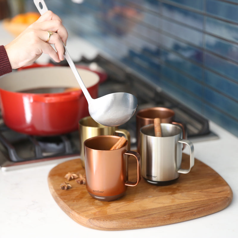 Ember Mug²: Metallic Collection filled with cinnamon sticks sitting on a wooden board while a person ladles mulled wine into each.
