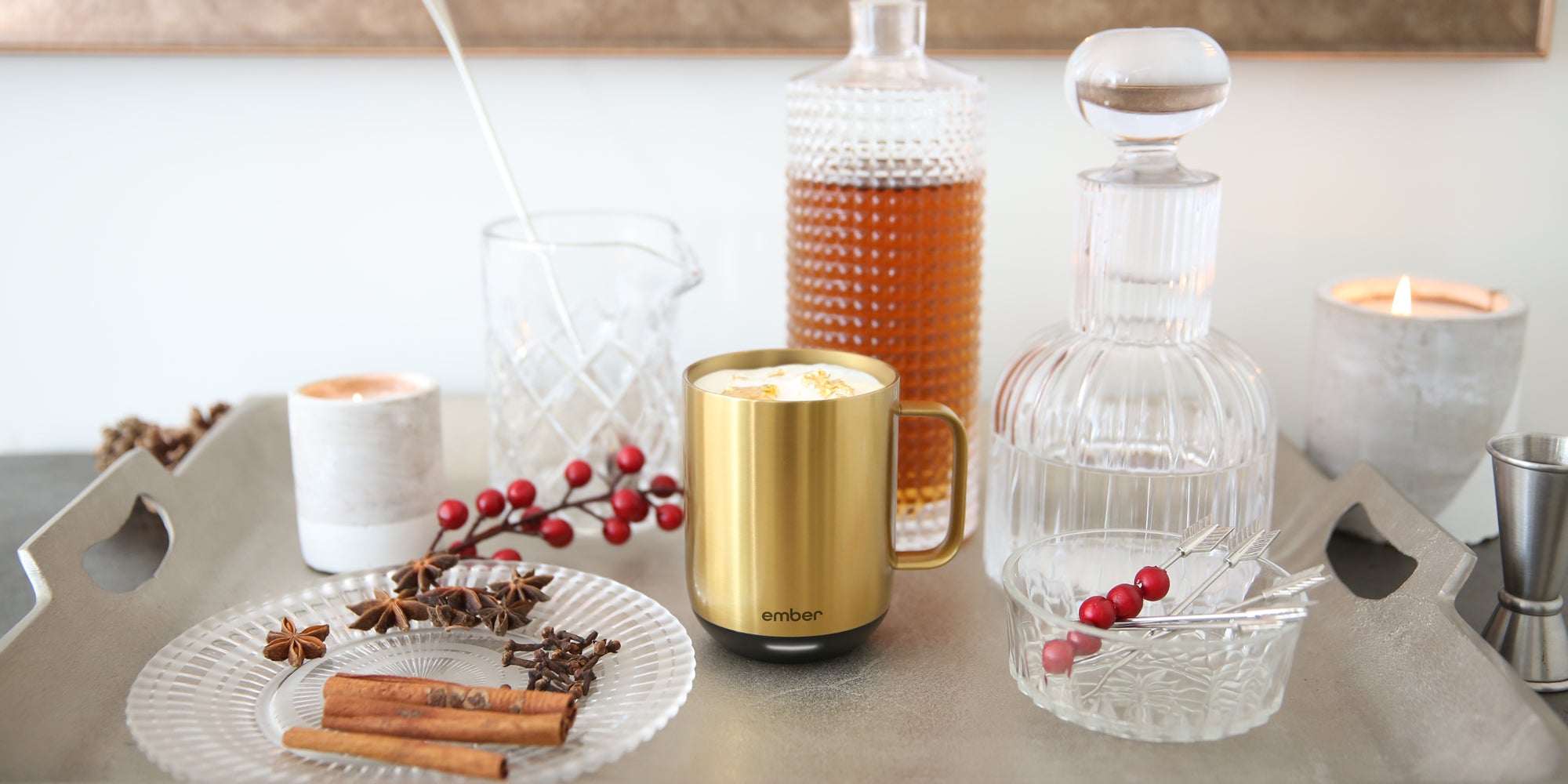 A gold Ember Mug² filled with a creamy warm beverage, surrounded with ornate glassware, cinnamon sticks, cloves, star anise, and cranberries.