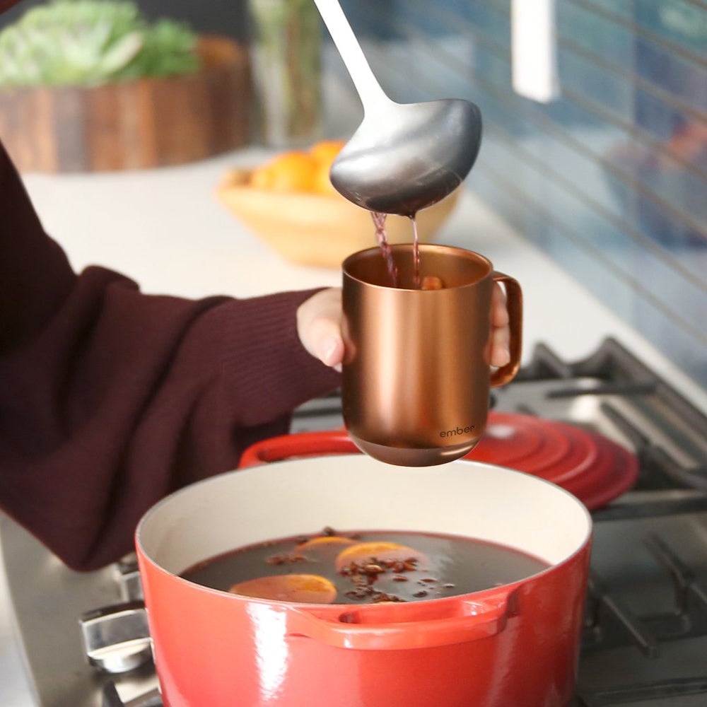 A person holding an Ember Mug² Copper Edition over a red pot of mulled wine while they ladle the beverage into the mug.