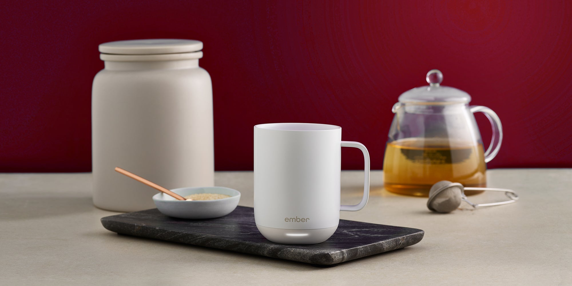 A white Ember Mug² sits on top of a dark marbled cutting board. There is a bowl of sugar to the left and a pot of tea to the right with a tea steeper. The background is dark red.